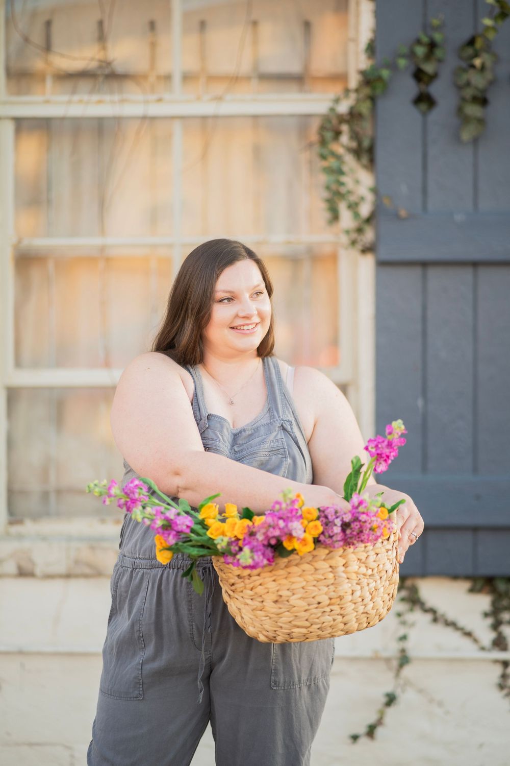 Cheerful woman in gray overalls holding wicker basket filled with bright spring flowers outdoors.
