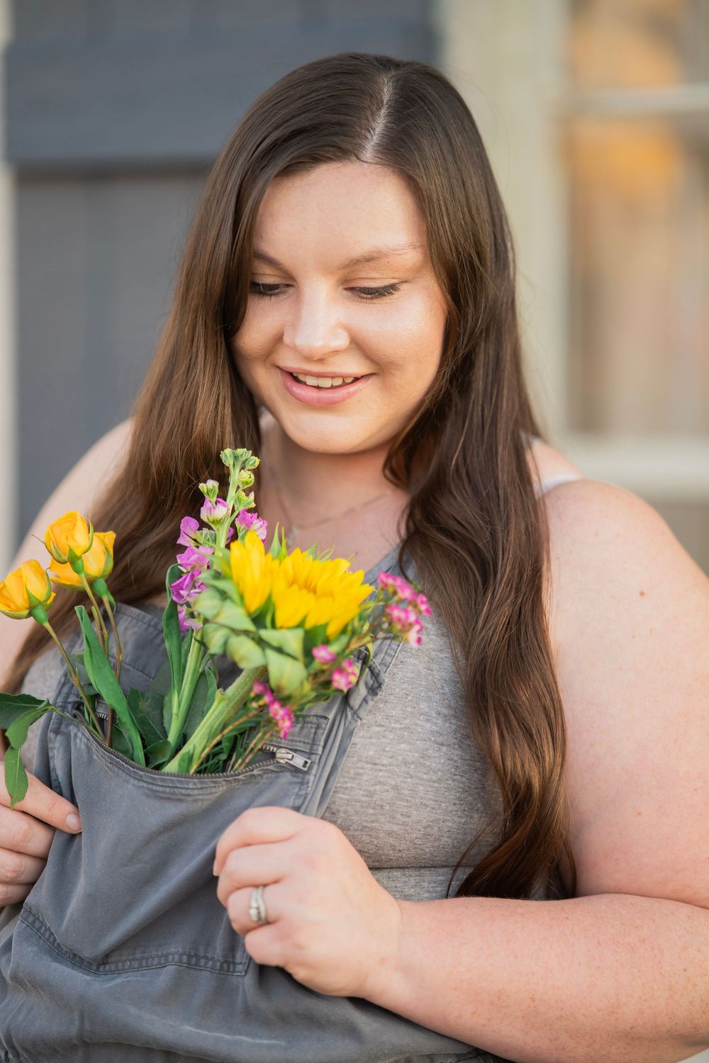Young woman in gray overalls smiling while holding yellow and pink flowers close to chest.