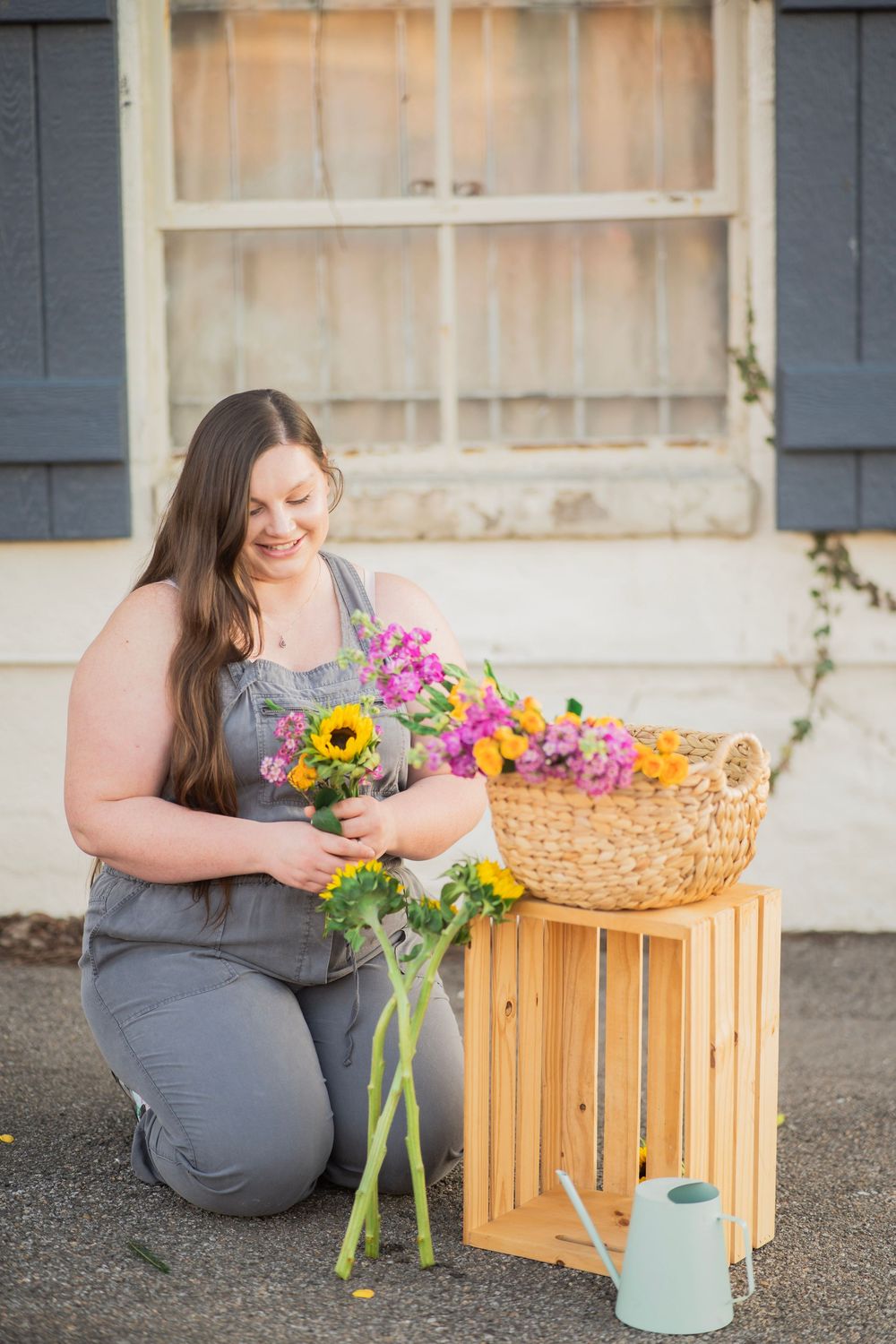 Woman in gray overalls kneeling beside wooden crate arranging sunflowers and colorful blooms.