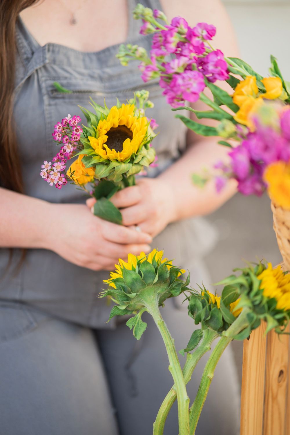 Close-up of woman in gray overalls arranging sunflowers and purple blooms from wicker basket.