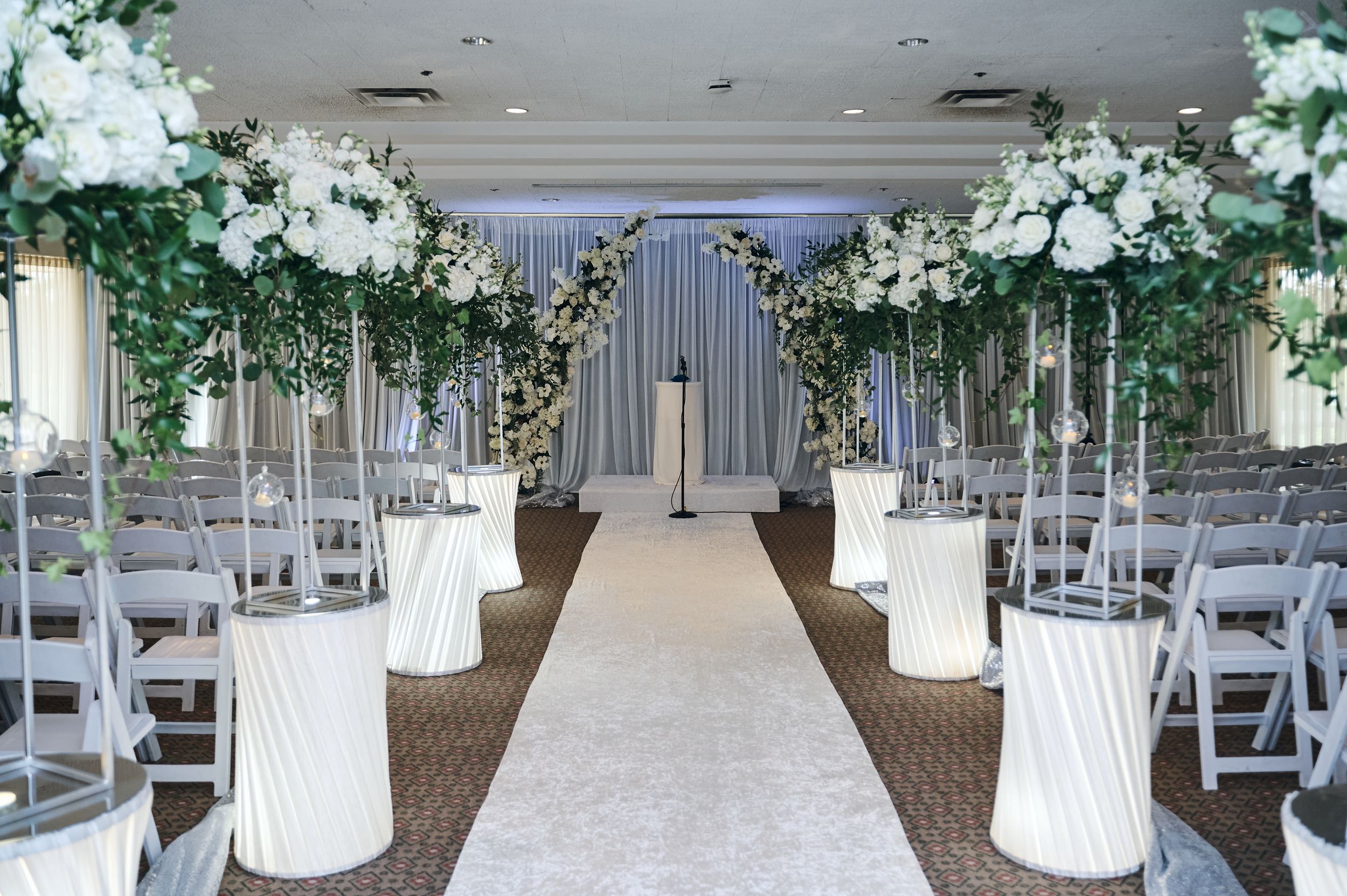 Elegant wedding ceremony aisle with white floral arch, tall white pedestals, and lush greenery in a decorated venue.