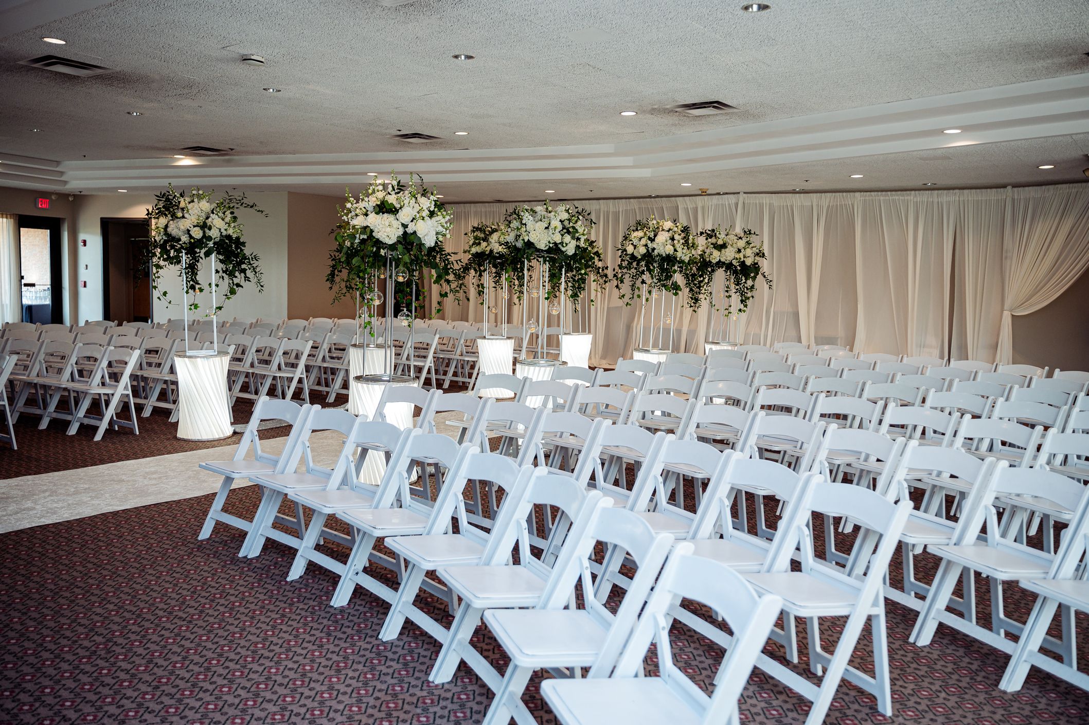 Elegant wedding ceremony setup with white folding chairs and floral arrangements in a decorated banquet hall.