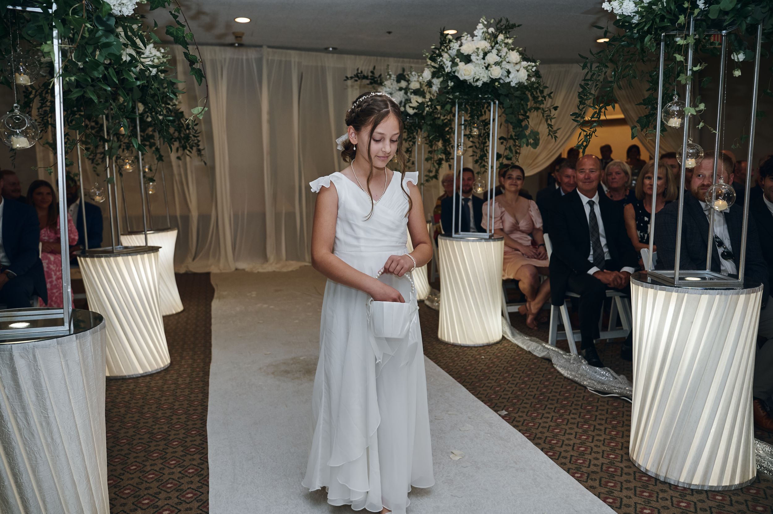 Bride in white gown walks down floral-decorated wedding aisle with seated guests watching during indoor ceremony.