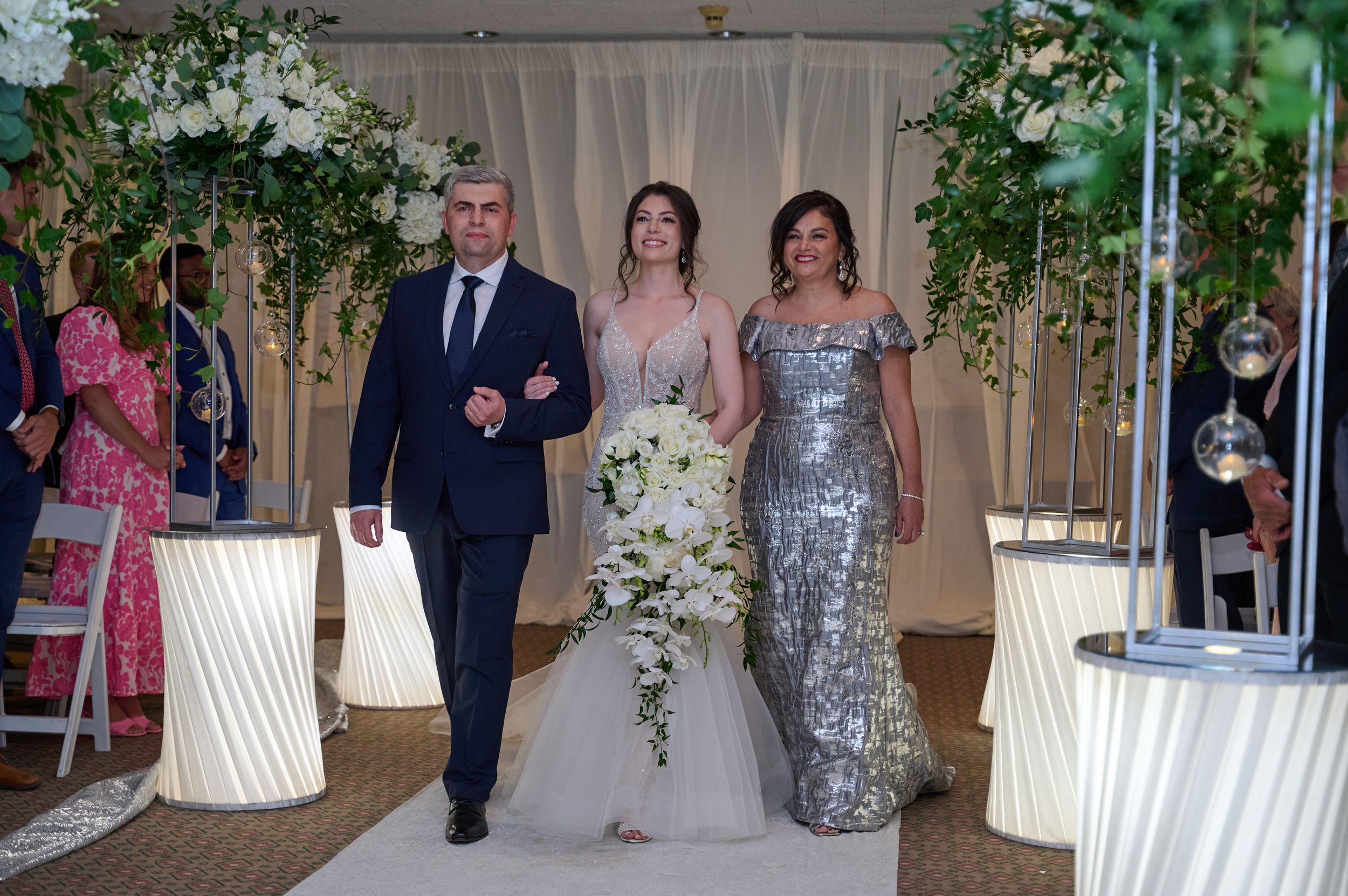 Bride walks down the aisle escorted by parents at elegant indoor wedding ceremony with floral arch.