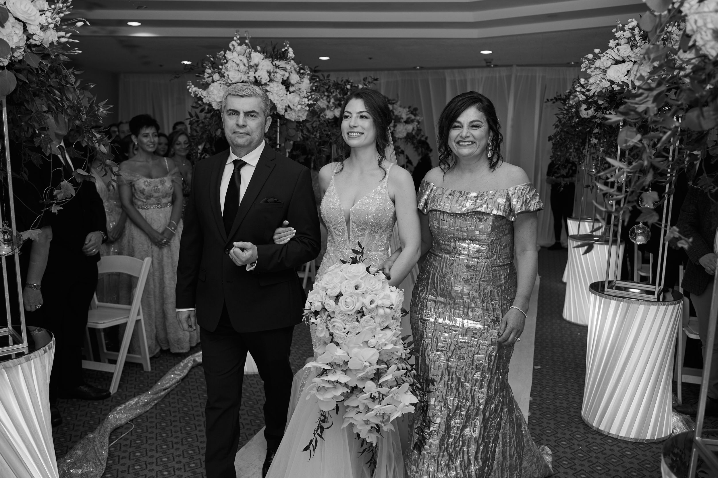 Black and white wedding photo of bride walking down aisle escorted by parents in elegant formal attire.