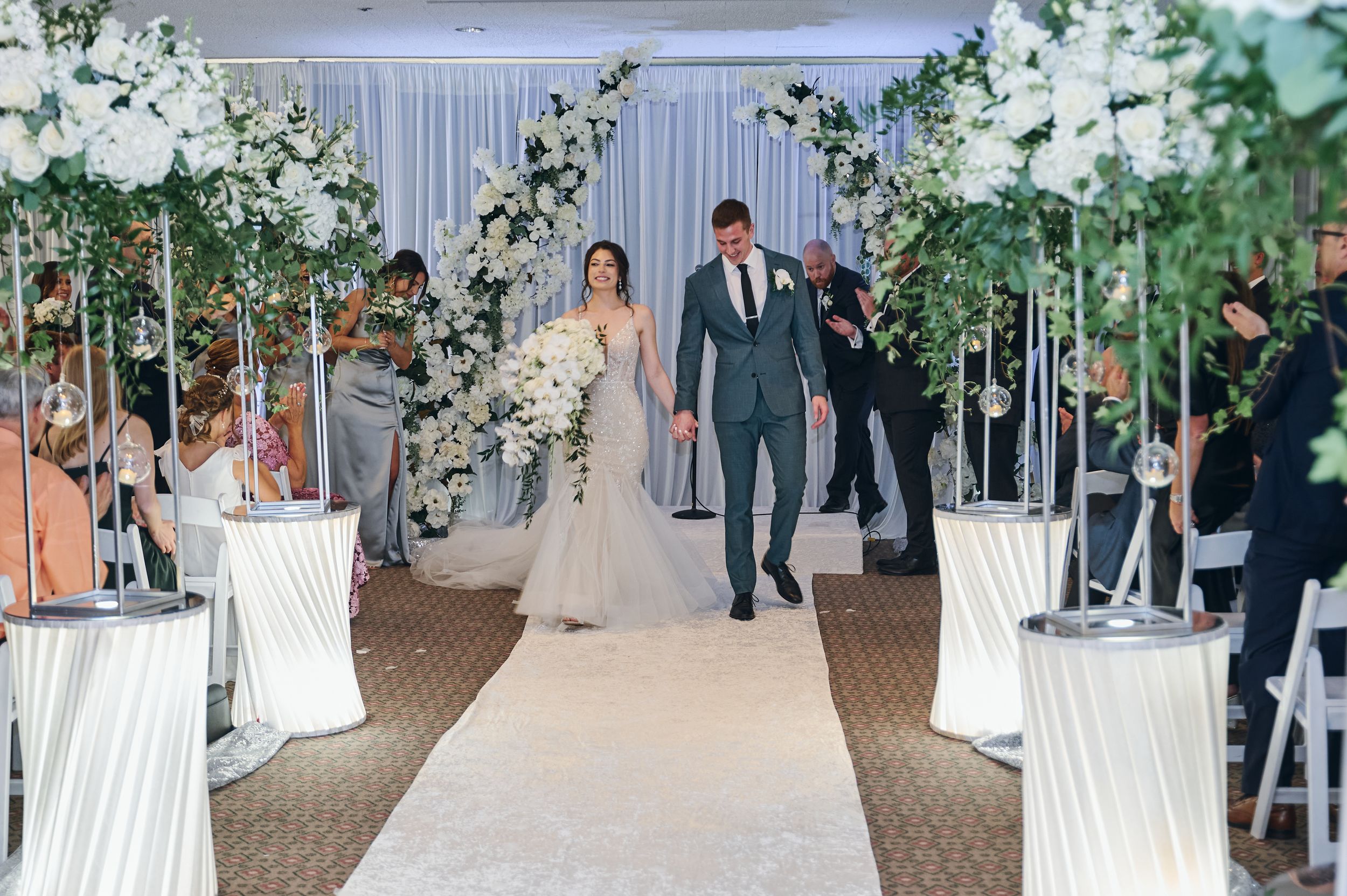 Bride and groom walk down floral-adorned aisle at elegant indoor wedding ceremony with white flowers and greenery.