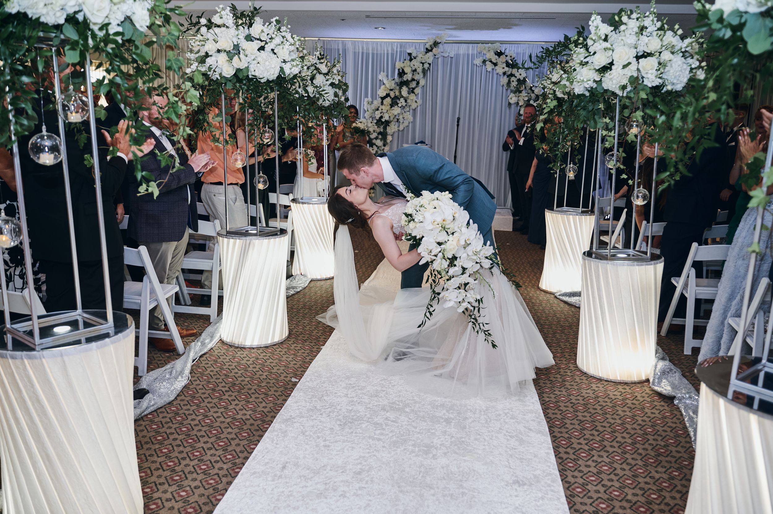 Bride and groom share a romantic kiss at indoor wedding ceremony with white floral arrangements and glowing pedestals.
