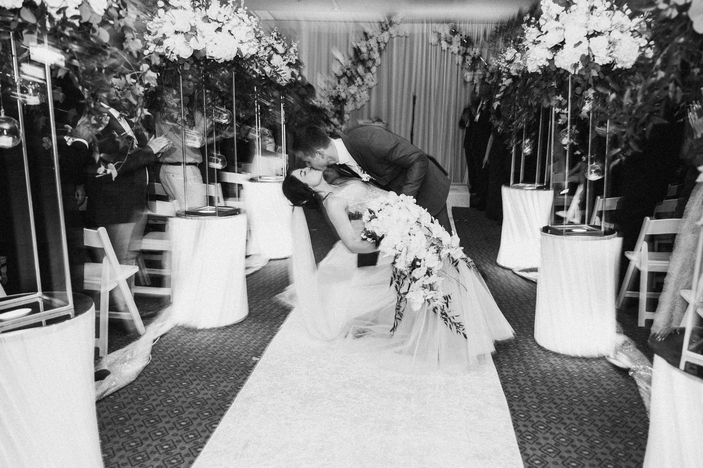 Black and white photo of groom dipping bride in wedding kiss at floral-decorated indoor ceremony aisle.