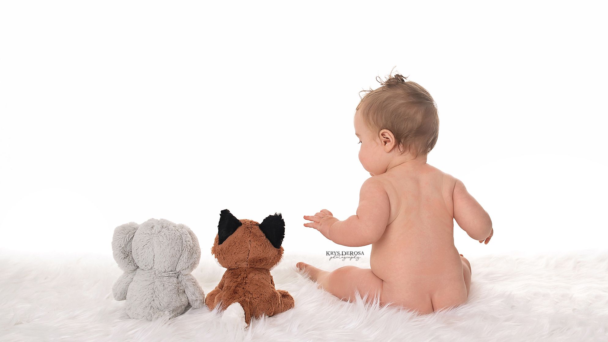 Baby sitting on white fur with a gray elephant and brown fox stuffed animals against a white background.