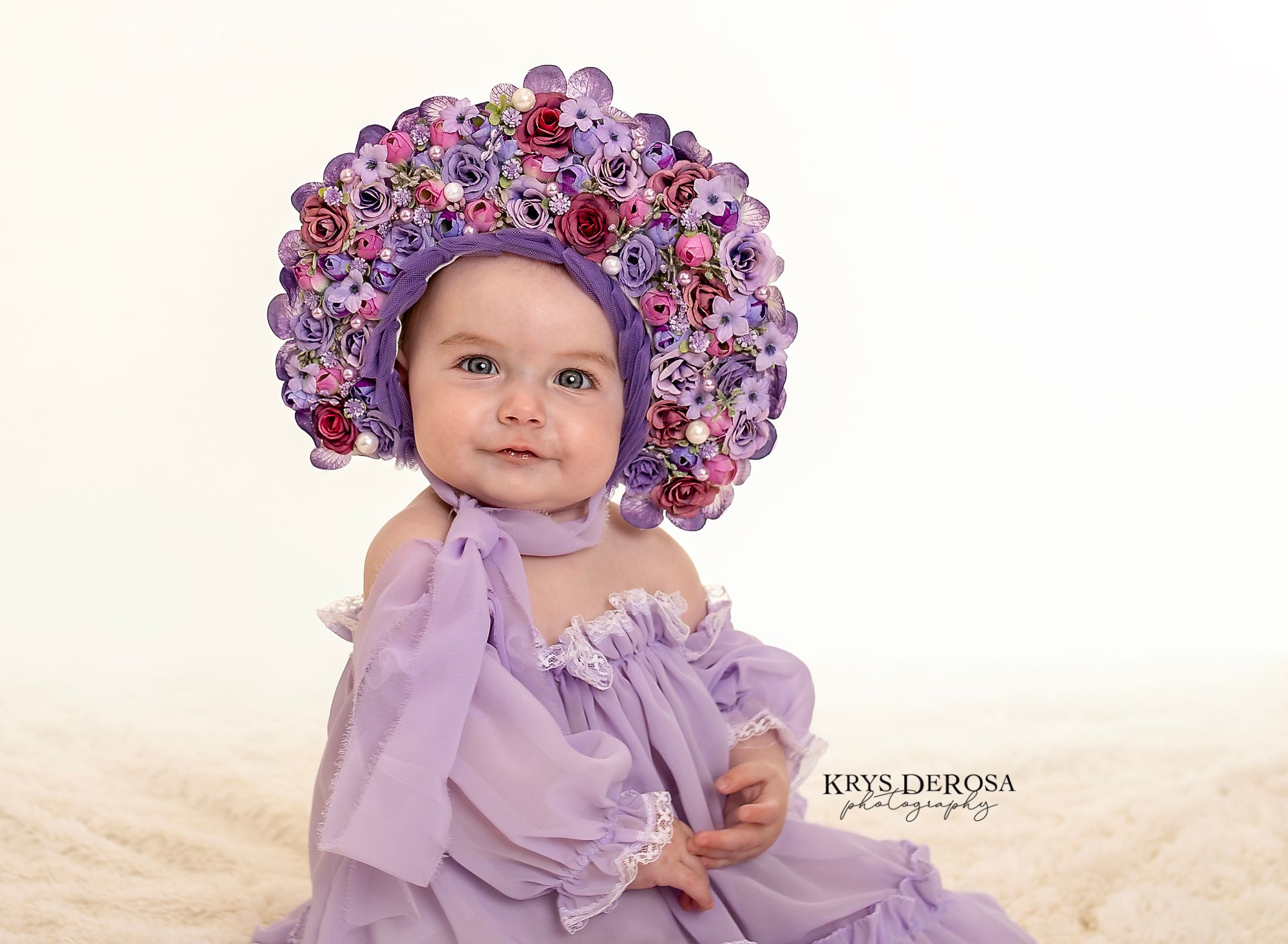 Baby in purple floral bonnet and lavender dress poses for professional portrait photography.