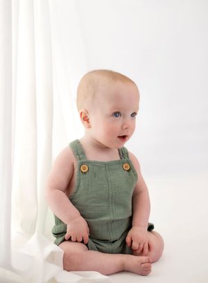 Baby in green linen overalls sitting on white backdrop, blue eyes, fair skin, wooden buttons detail.