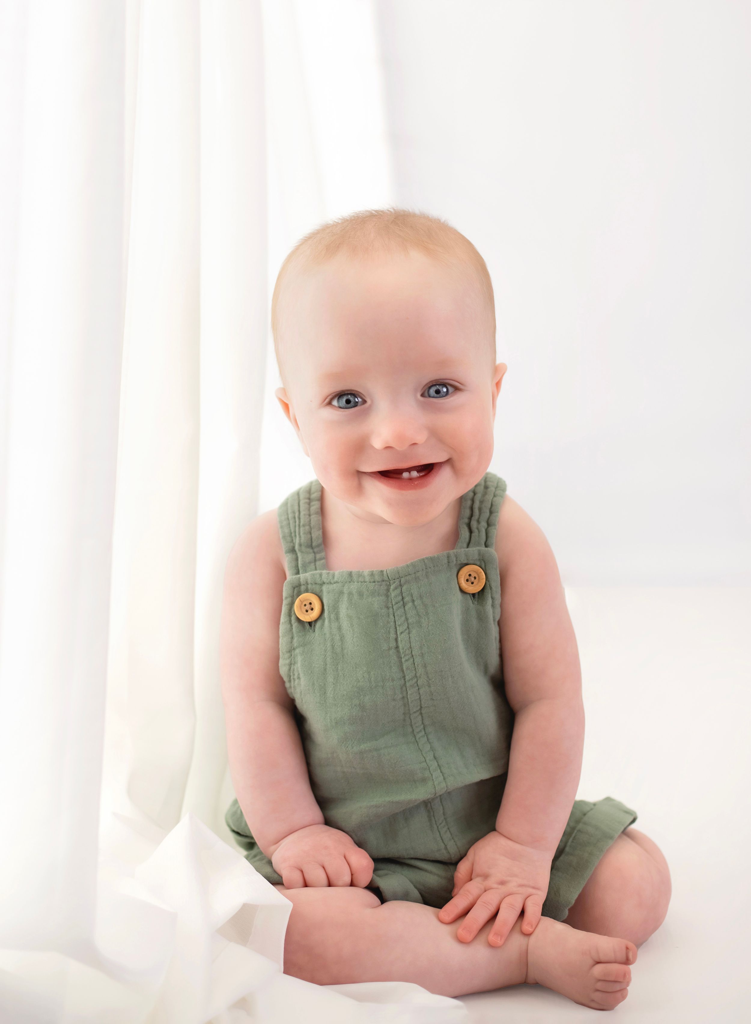 Happy baby boy in sage green button overalls smiling brightly in white studio.