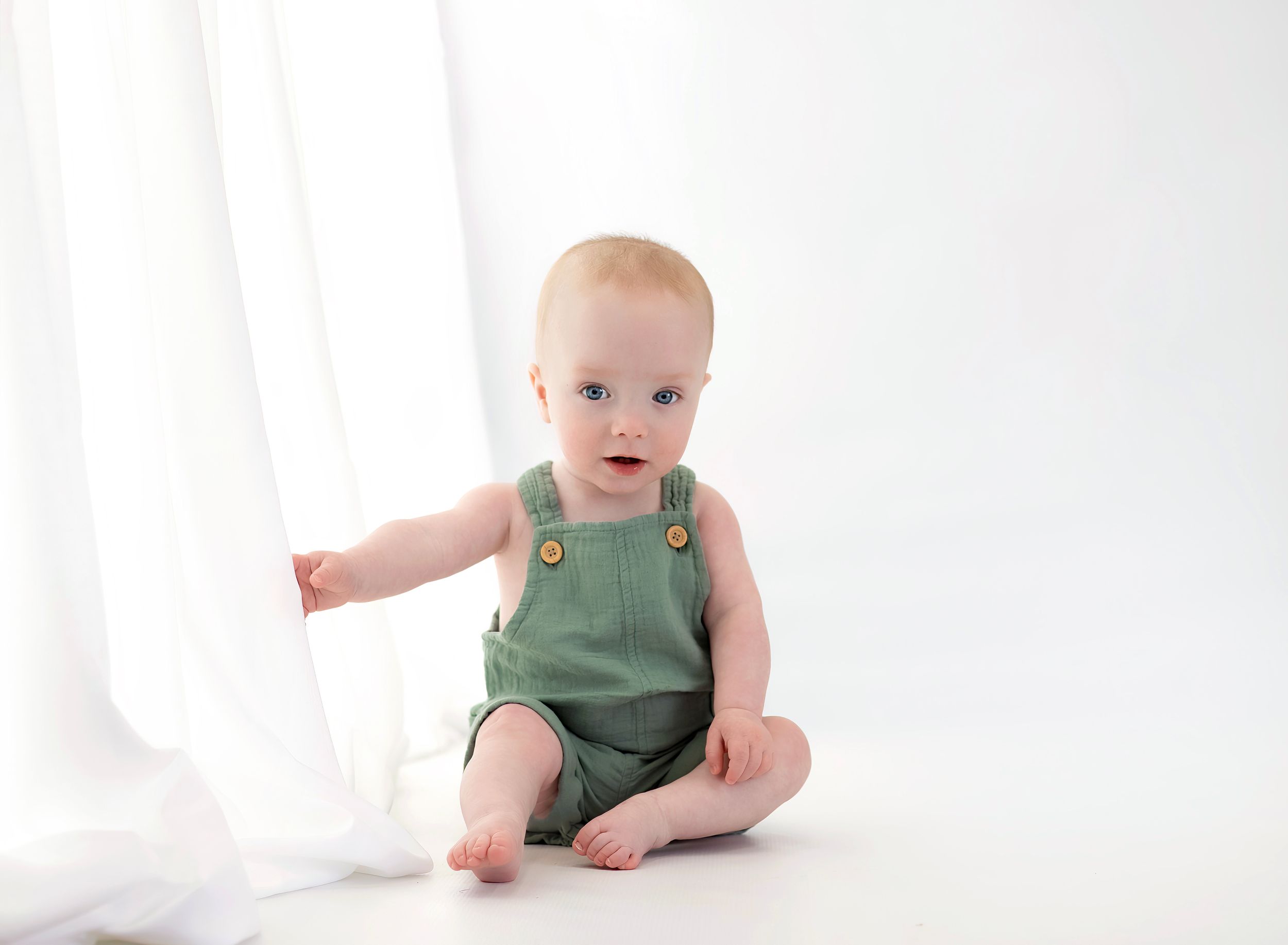 Adorable baby in green linen romper sitting on white background, reaching toward sheer curtain.