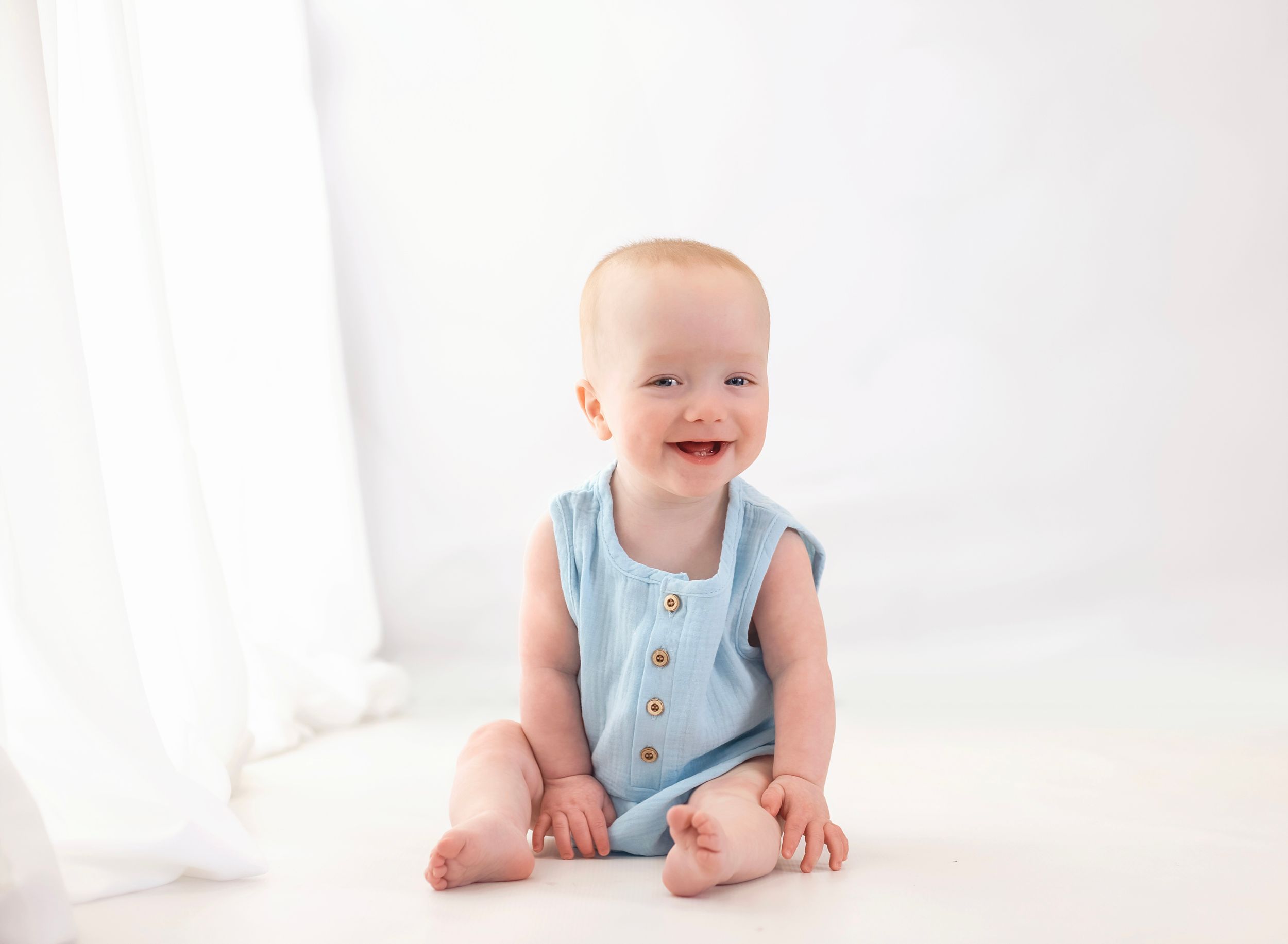 Happy baby in blue romper sitting and smiling on white background.