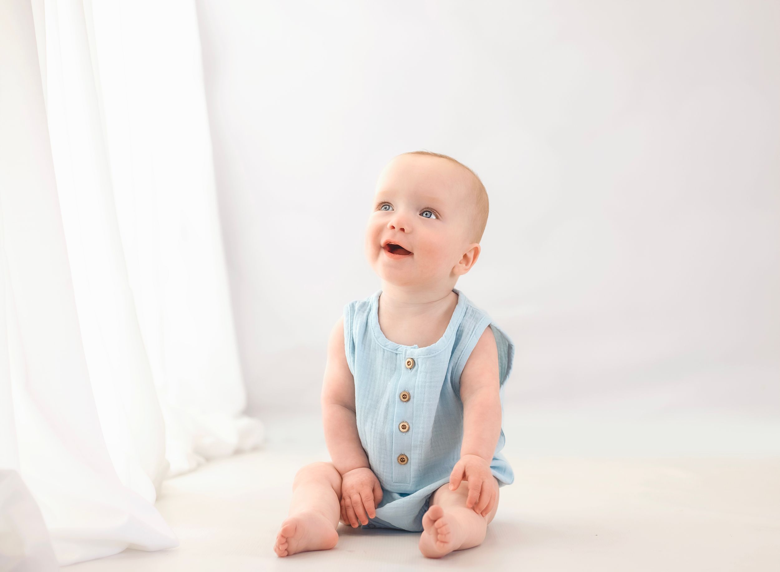 Adorable baby in light blue romper sitting and looking up in bright white studio.