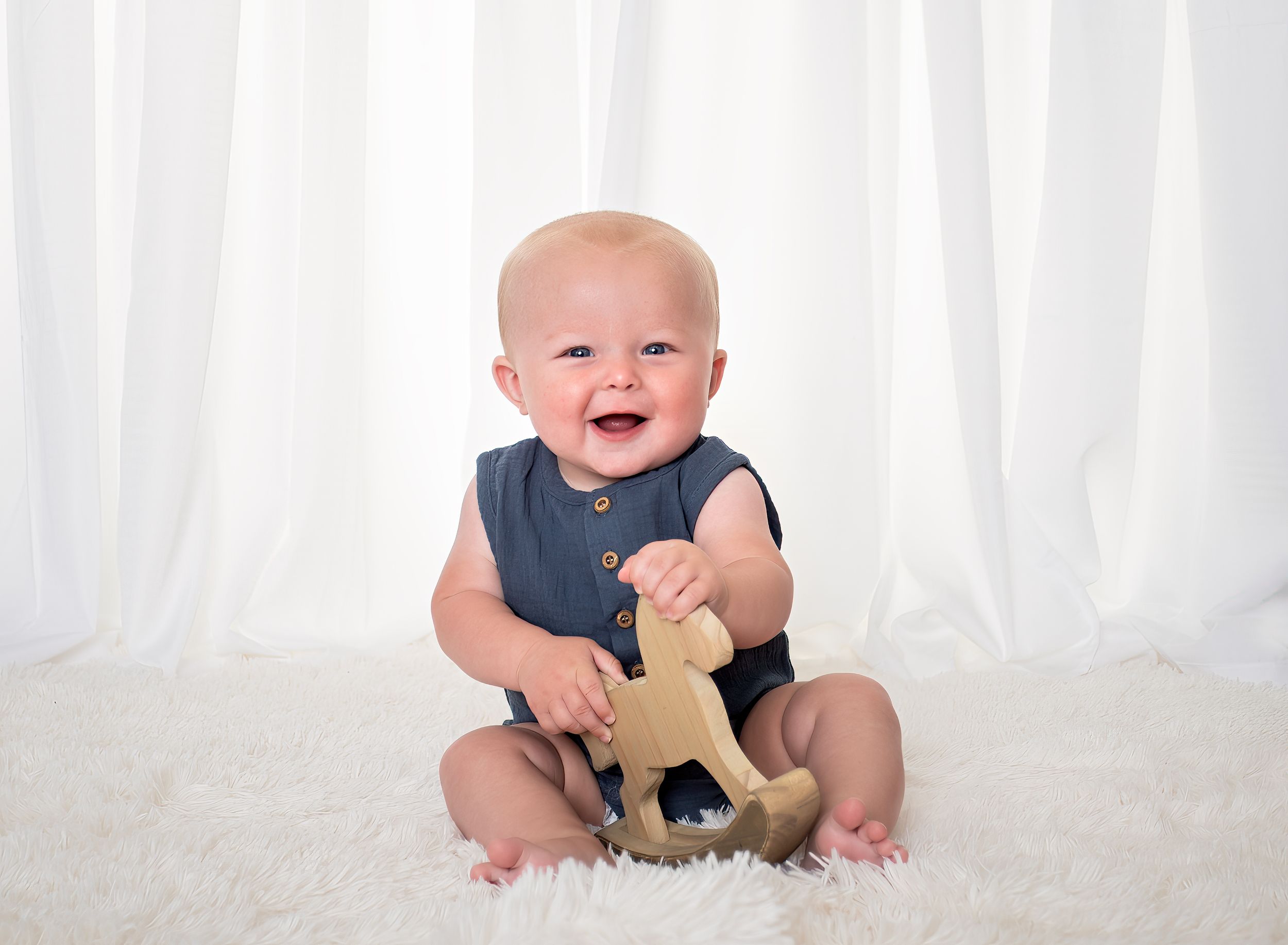 Smiling baby in navy romper sitting on white fur rug holding a wooden rocking horse toy.