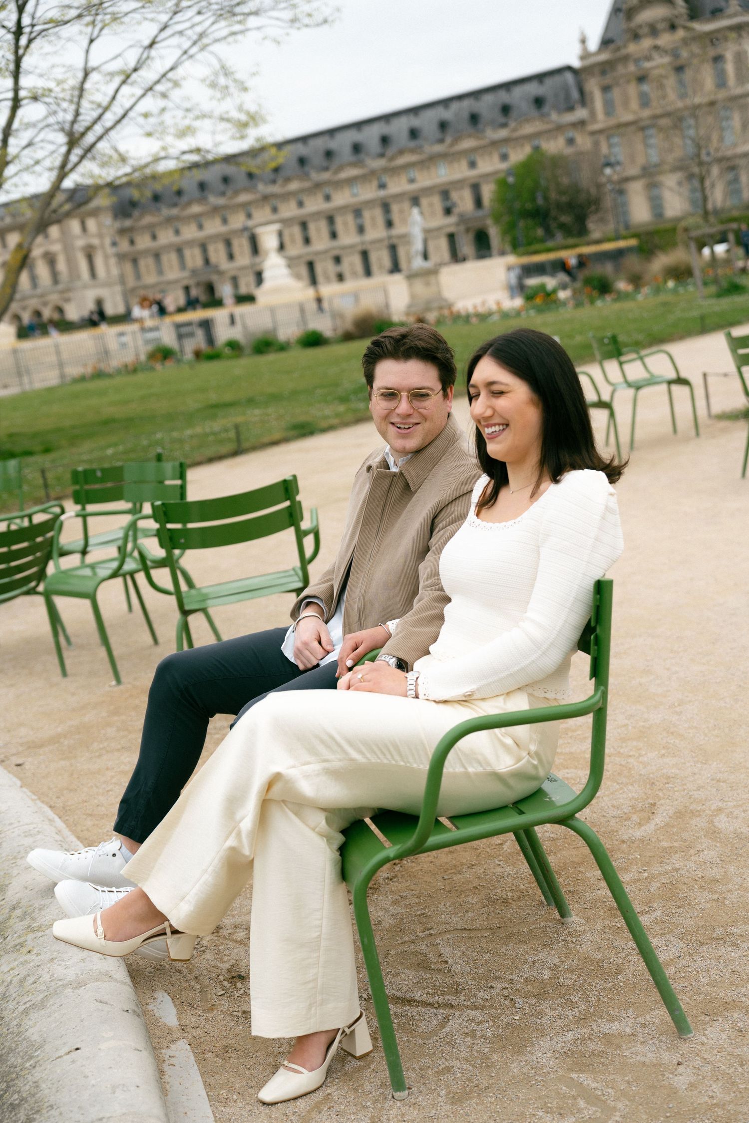 A couple smiles while sitting on green chairs in the Tuileries Garden, Paris, with the Louvre visible in the background. Paris editorial