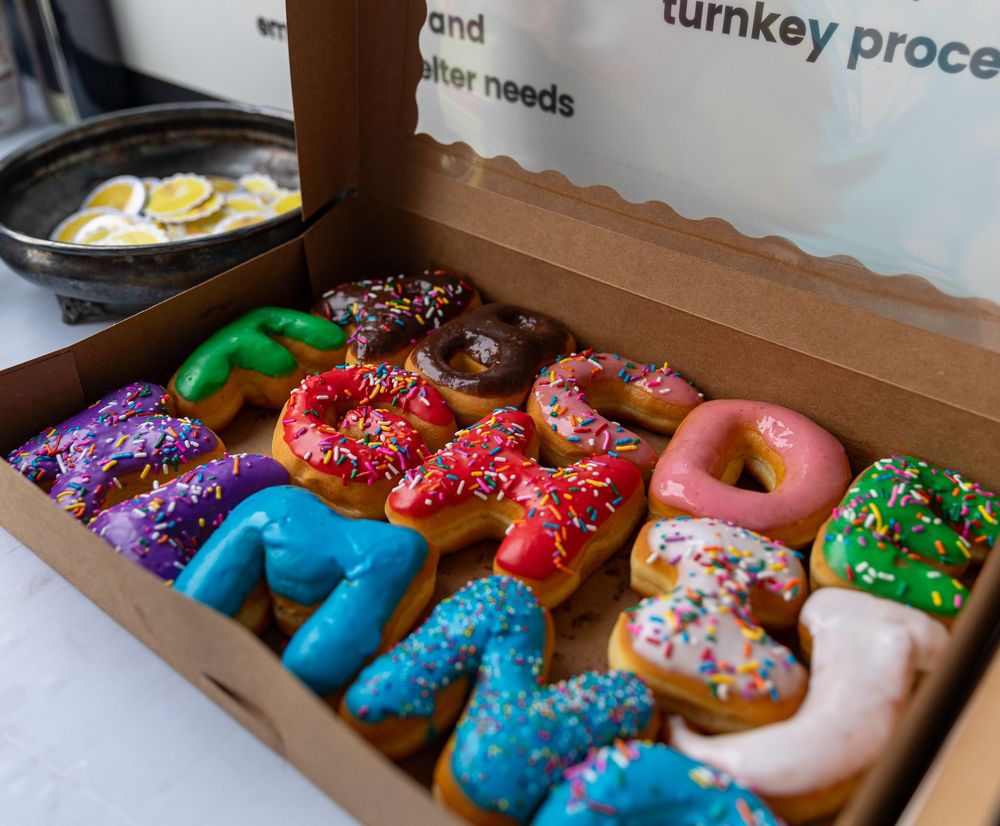 A box of colorful letter-shaped donuts with sprinkles spelling a word, displayed in a cardboard bakery box.