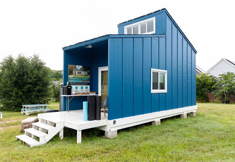 A small blue tiny house with white deck and stairs on a green lawn.