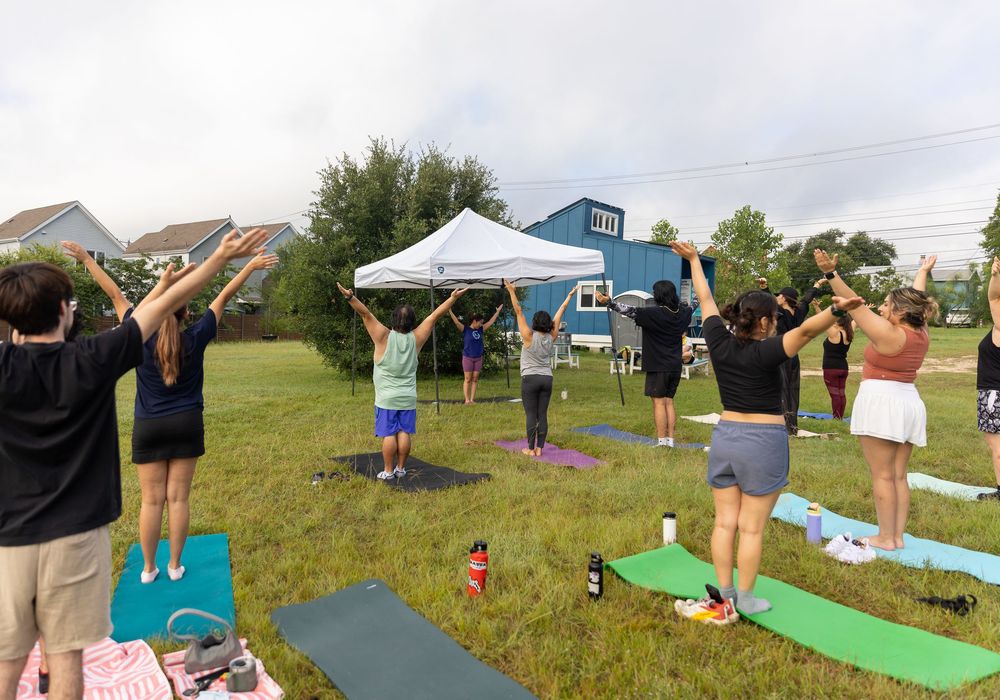 Group outdoor yoga class on green grass with participants raising arms near a white canopy tent on a cloudy day.