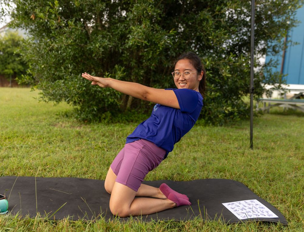 Woman smiling in yoga pose on black mat outdoors on green grass.