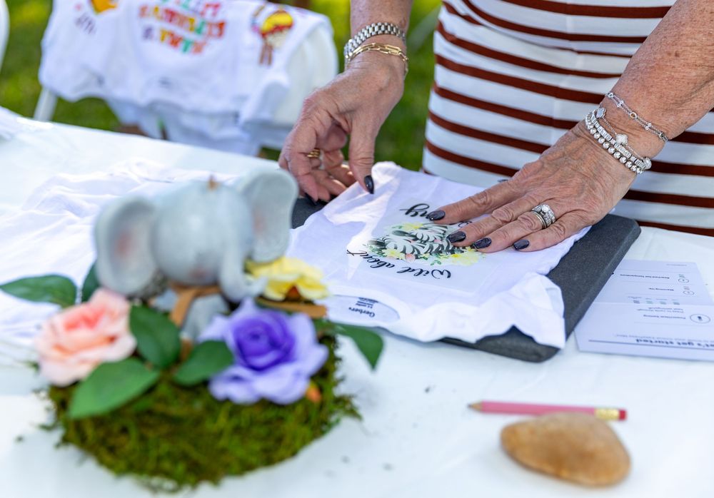A woman decorates a baby shower gift bag near a floral centerpiece with a gray elephant figurine on a white table.