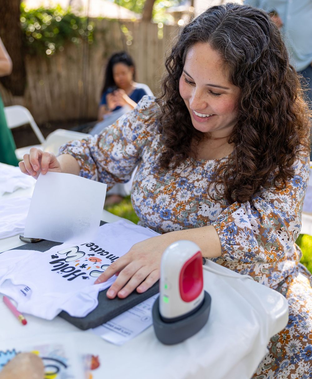 Woman smiling while applying heat transfer vinyl design to a onesie at an outdoor crafting event.