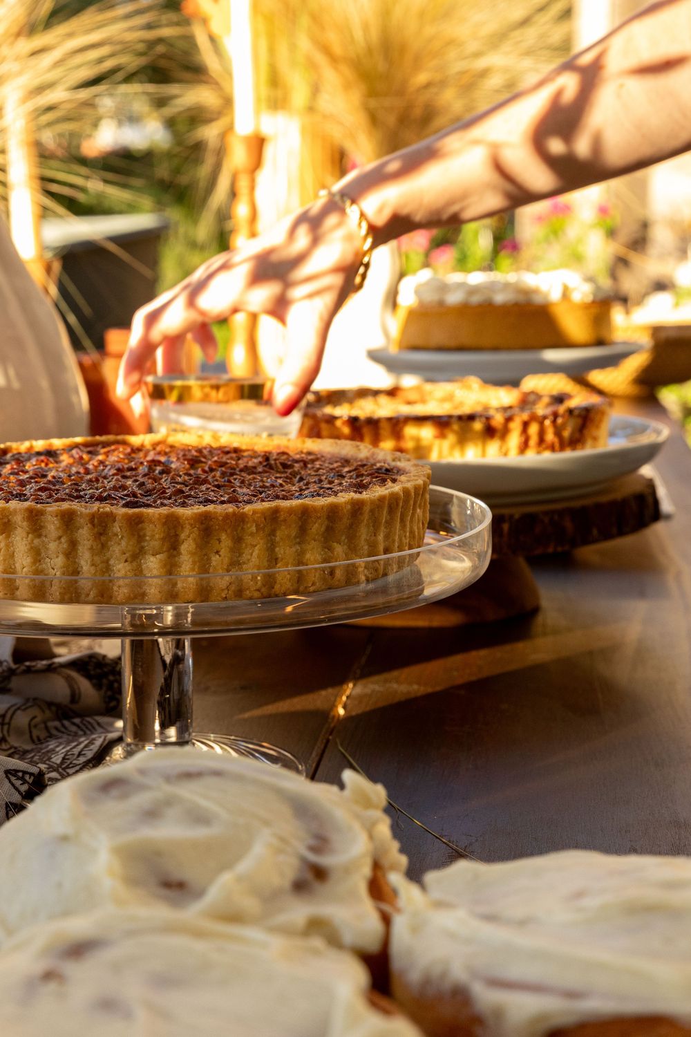 A hand reaches for a pecan tart on a glass stand surrounded by assorted pies on a rustic table in warm golden light.