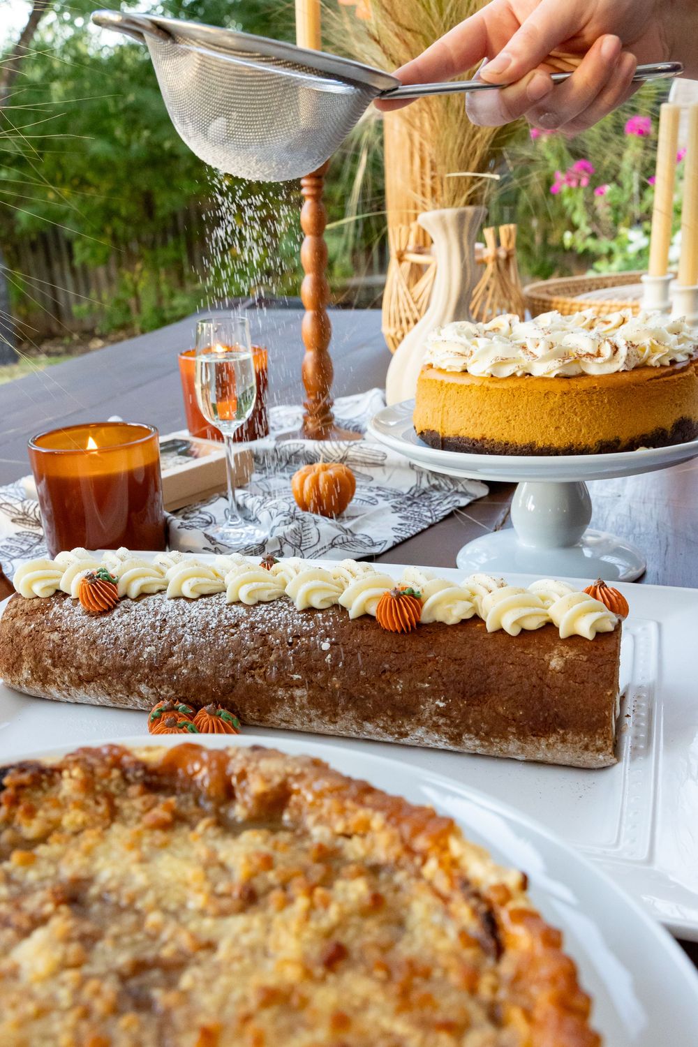 Hand dusting powdered sugar over fall dessert table with pumpkin cheesecake, log cake, and pie outdoors.