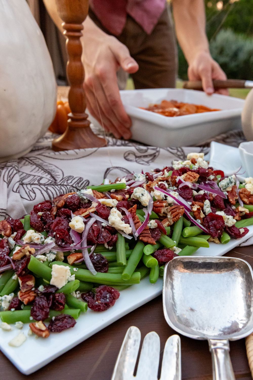 A colorful green bean salad with cranberries, red onion, pecans, and blue cheese on a white platter at a dinner table.