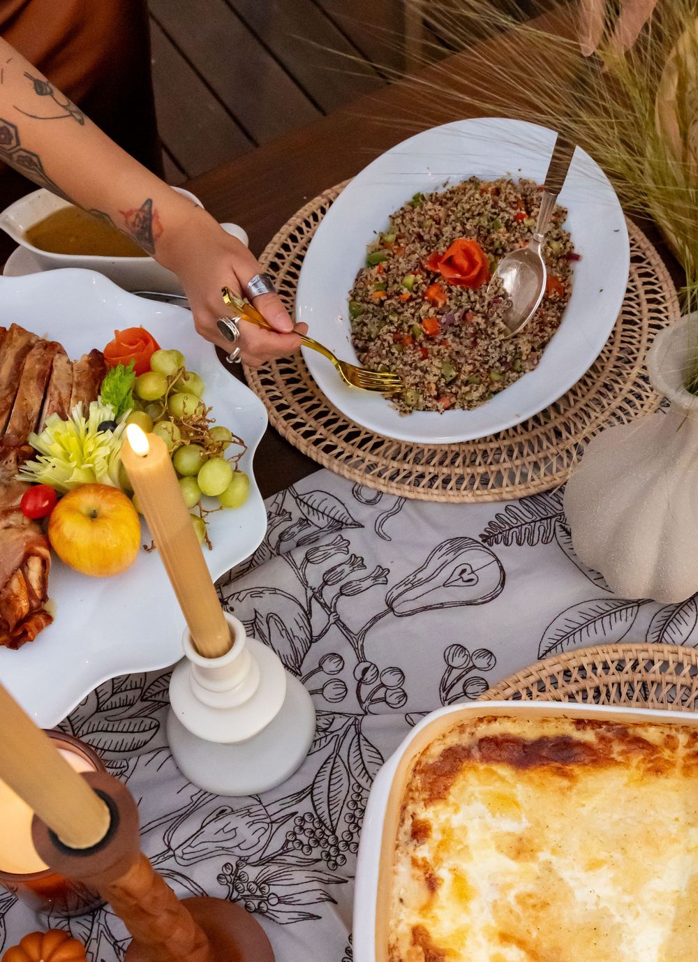 A tattooed hand serves grain salad at a boho Thanksgiving table with lasagna, roasted meat, candles, and floral tablecloth.