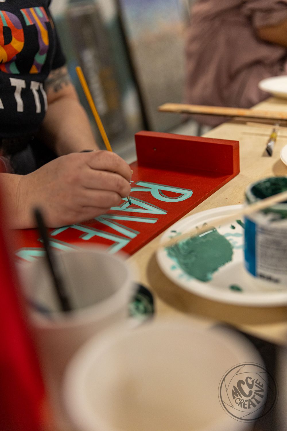 Artist painting teal letters on a red sign at a creative workshop, with paint palette and brushes visible.