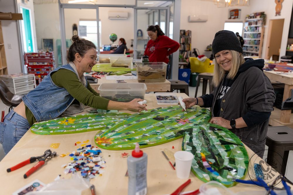 Two women smile while working on a colorful mosaic art project together at a table in a bright studio workshop.
