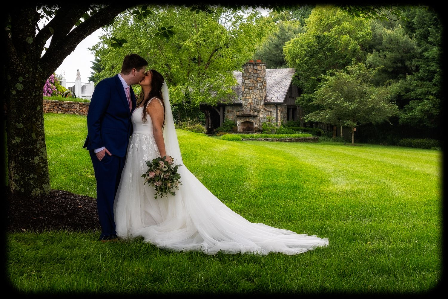 Bride and groom share a romantic kiss under a tree on lush green lawn with stone chapel in background.