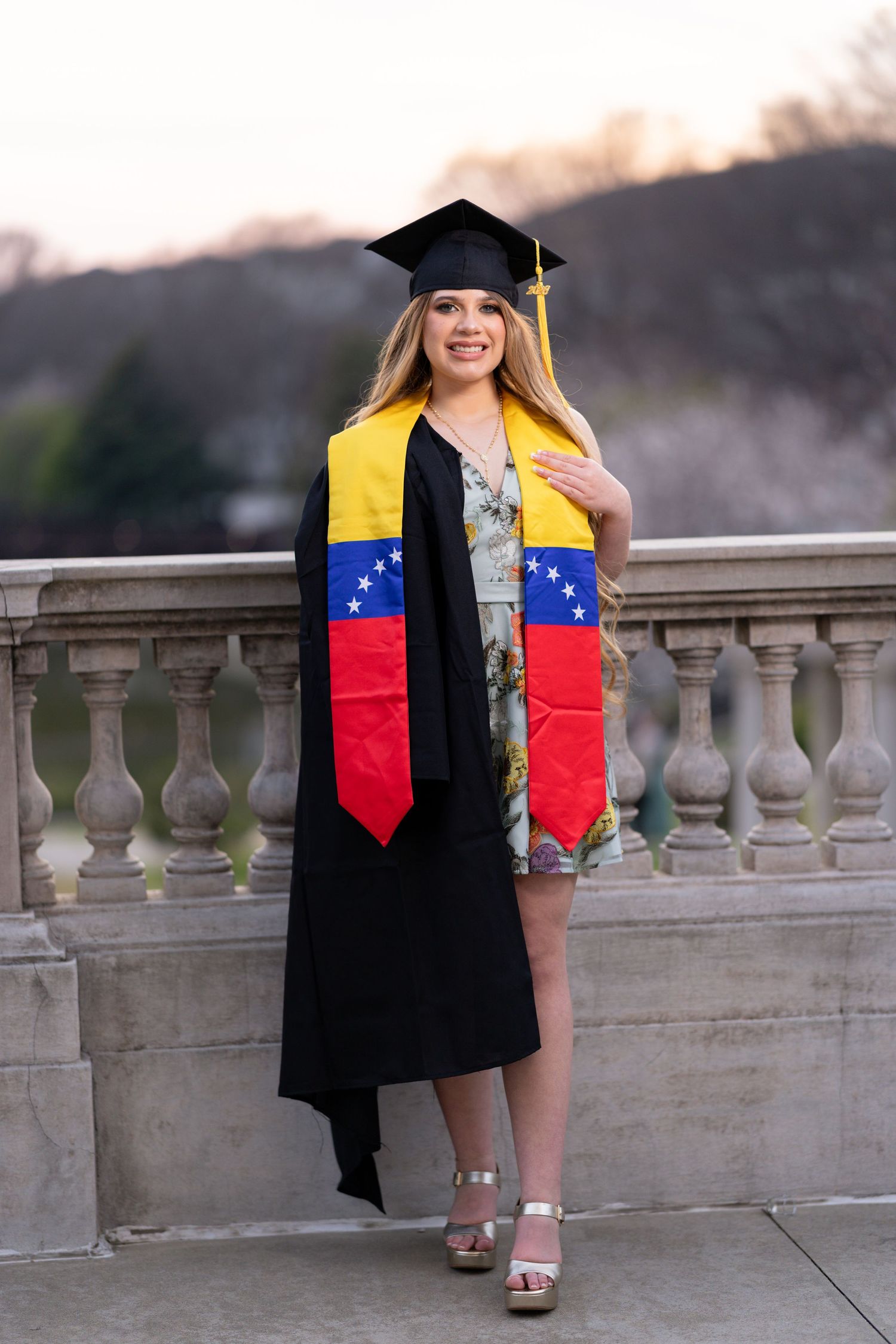 Smiling graduate in black cap and gown wearing Venezuelan flag sash, posing outdoors on a stone balcony.