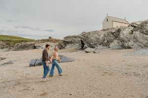 Couple walking hand in hand on a sandy beach beside rocky cliffs with a white cottage in the background.