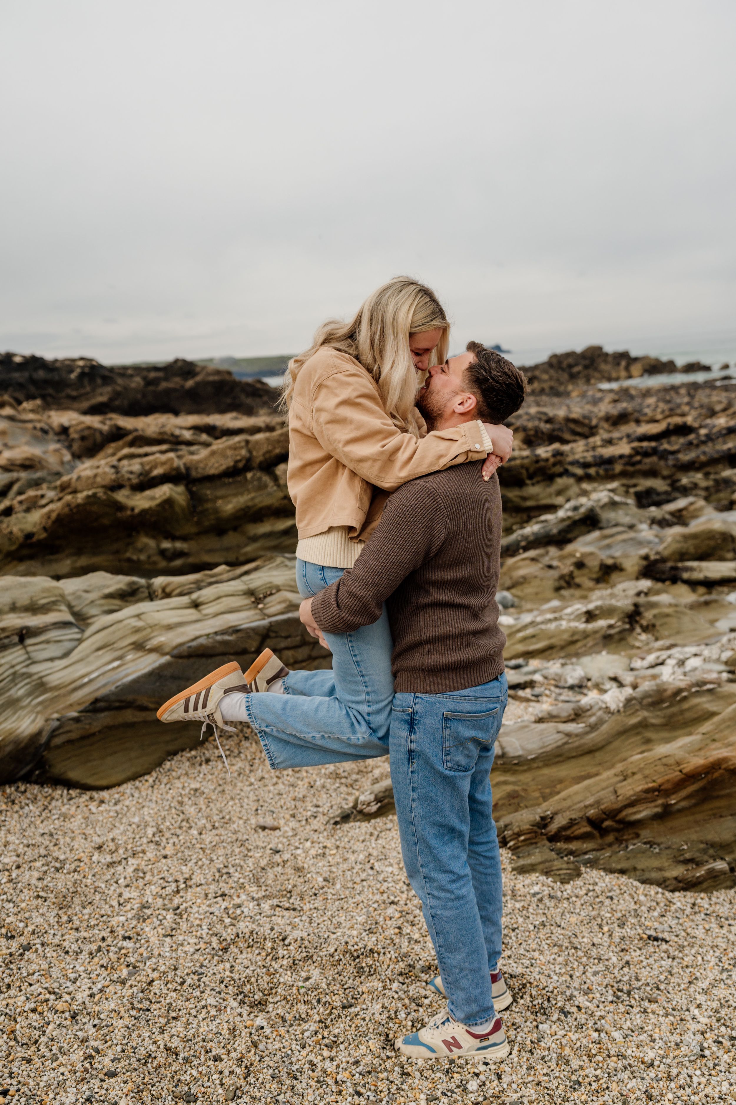 A couple shares a romantic kiss on a rocky beach, with the man lifting the woman in his arms.