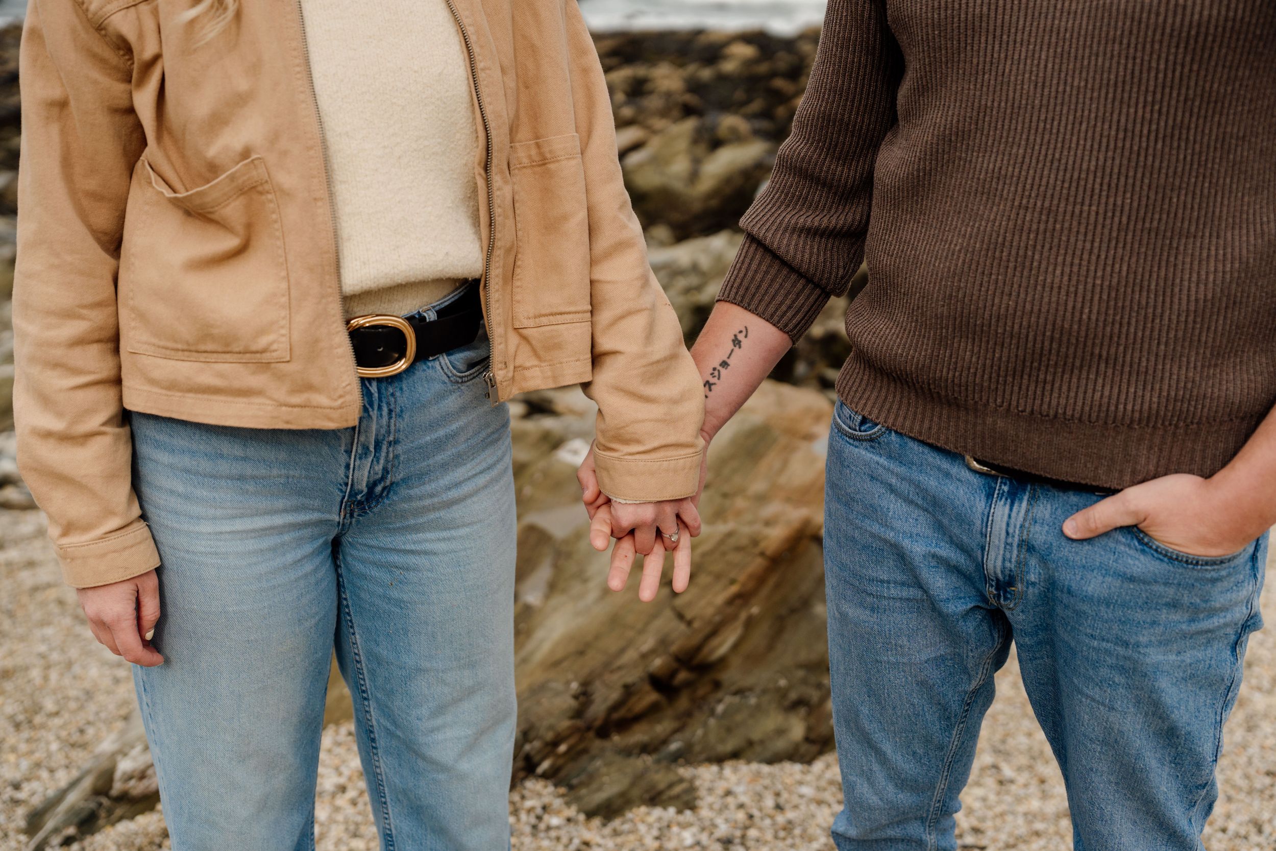 Couple holding hands on a rocky beach, wearing casual jeans and sweaters, with a visible tattoo on one wrist.