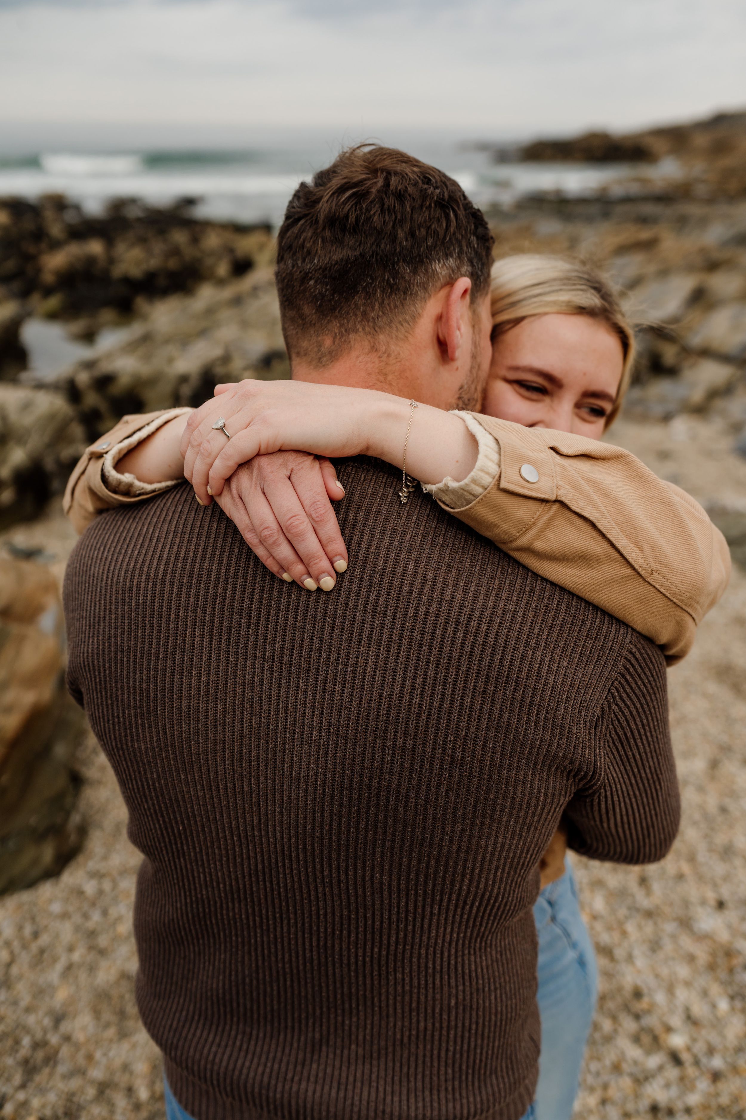 A couple embraces on a rocky beach, the woman smiling as she wraps her arms around the man in a brown sweater.
