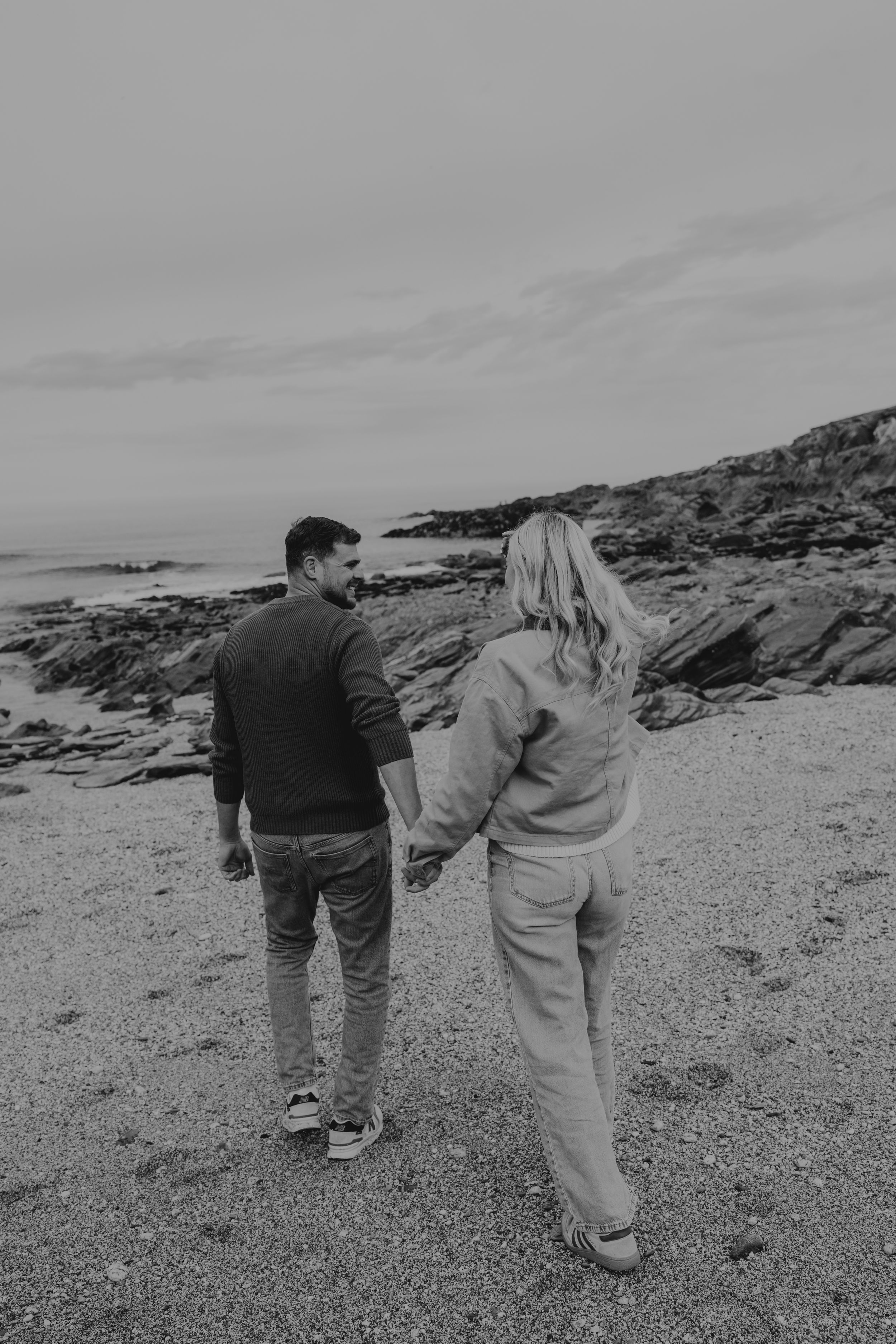 Black and white photo of a couple holding hands on a rocky beach, walking toward the ocean on an overcast day.