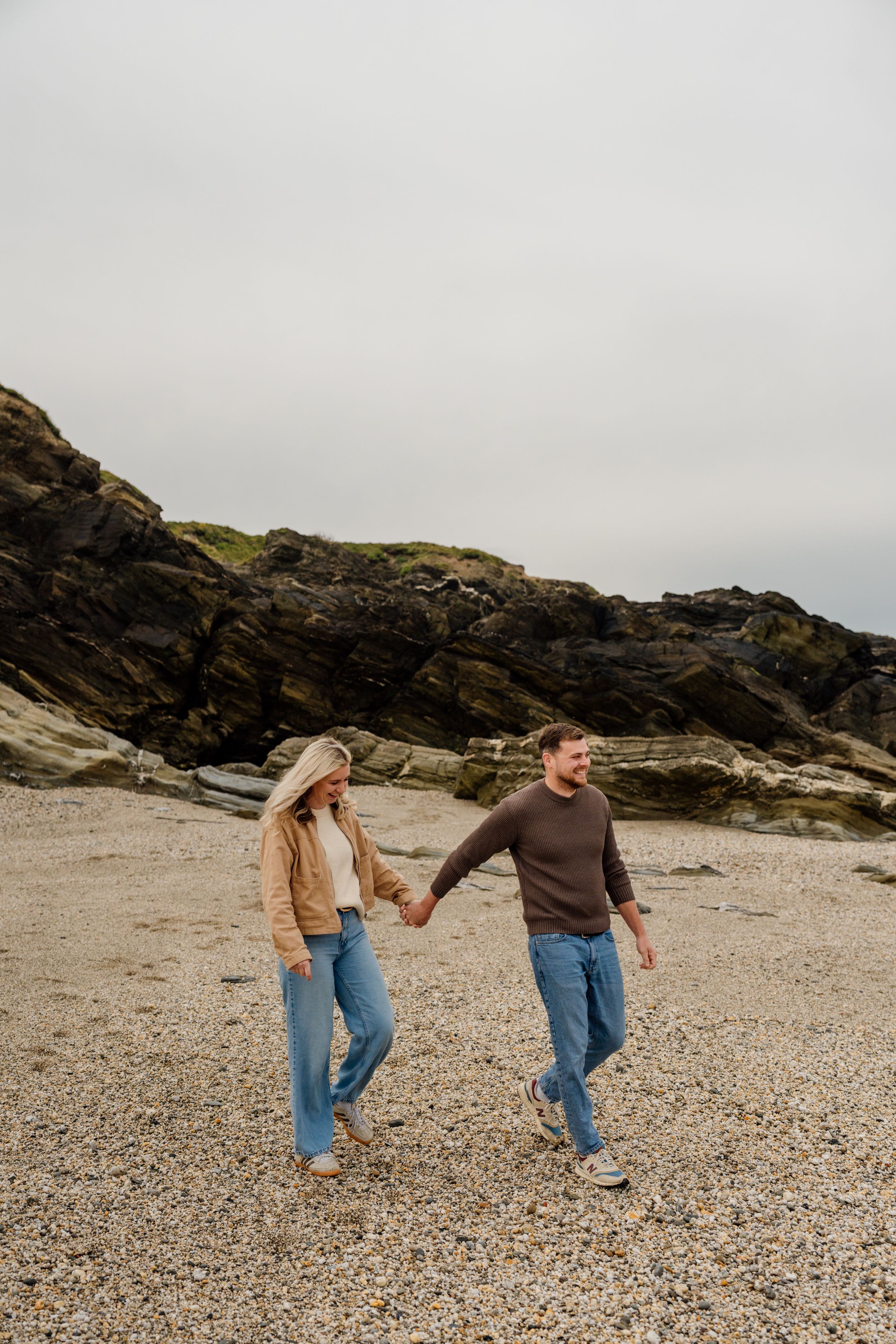 Couple holding hands walking on a rocky beach, wearing casual jeans and jackets with dramatic cliffs behind them.