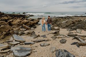 Couple holding hands on rocky beach with ocean waves and cloudy sky in background.