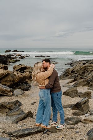 Couple embracing and kissing on a rocky beach with ocean waves in the background on a cloudy day.