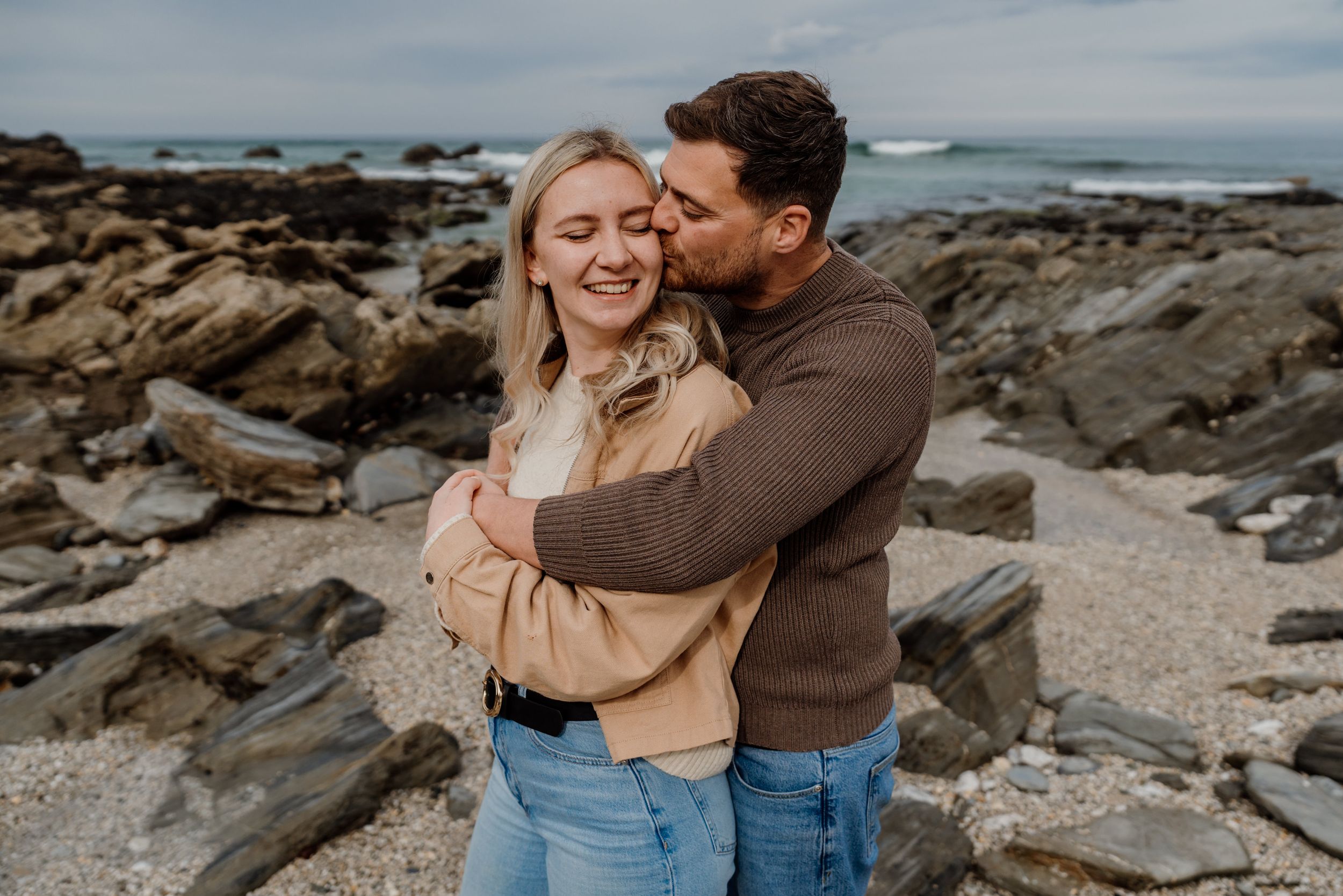 Couple embracing on rocky beach, woman smiling with arms crossed, man kissing her cheek, overcast sky backdrop.