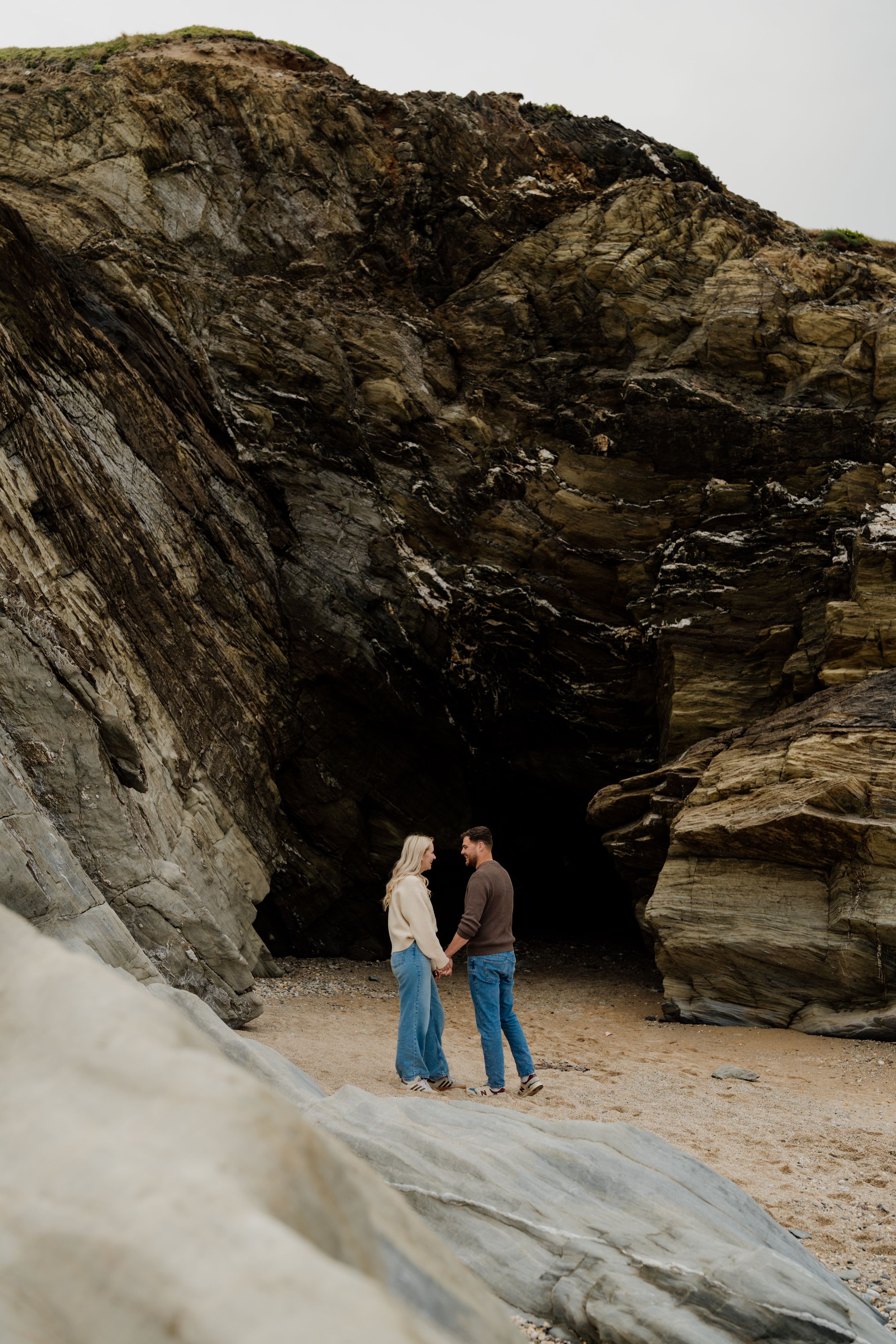 Couple holding hands on sandy beach in front of dramatic rocky cave cliffs on an overcast day.
