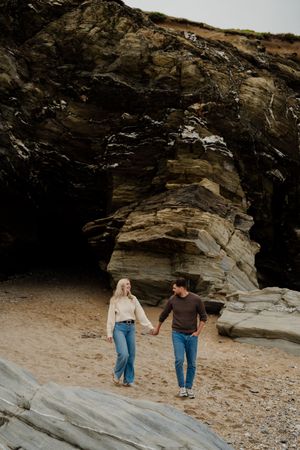 Couple holding hands walking on sandy beach with dramatic layered rock formations in background.