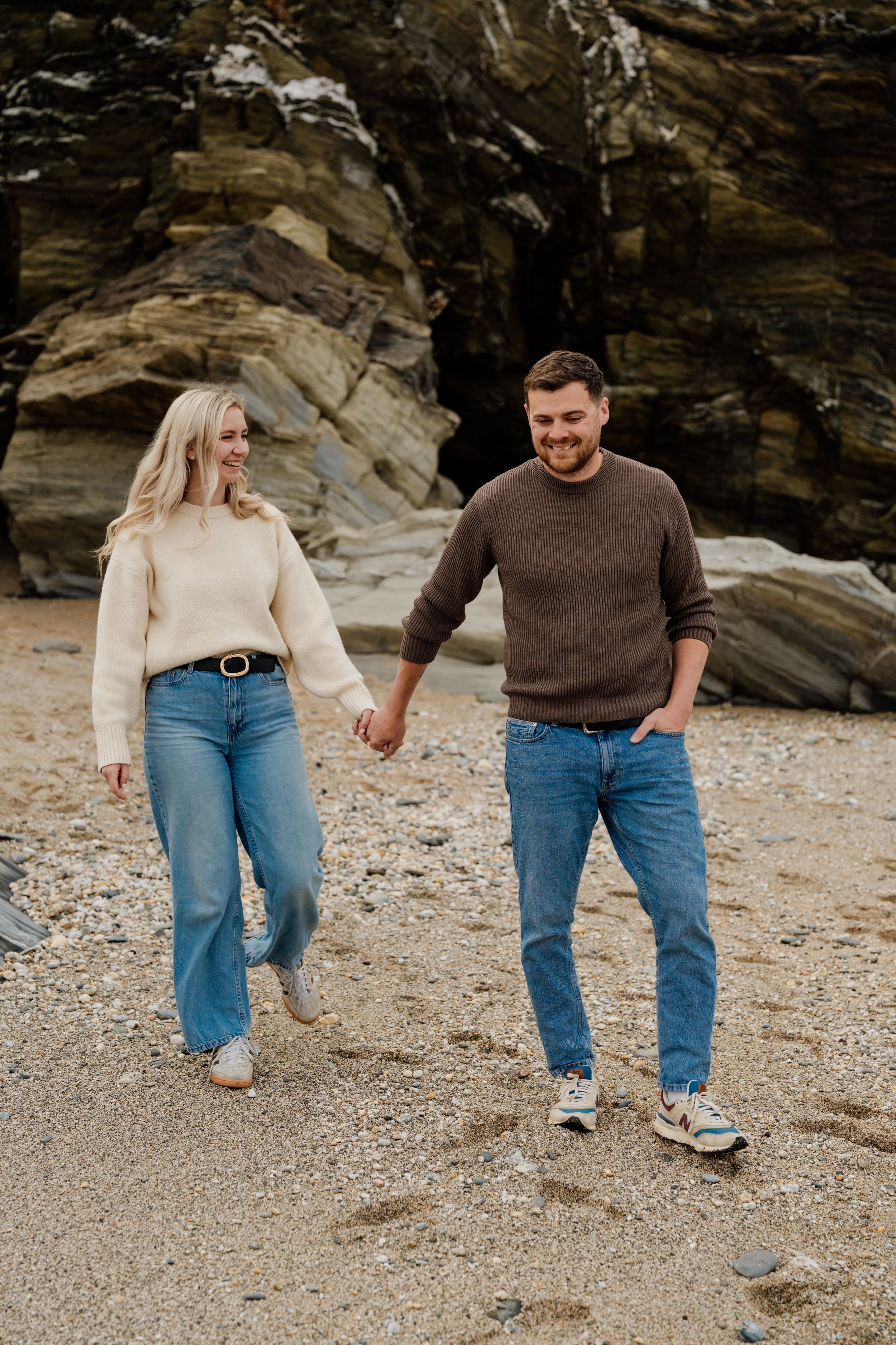A smiling couple holds hands while walking on a rocky beach, wearing casual sweaters and jeans.