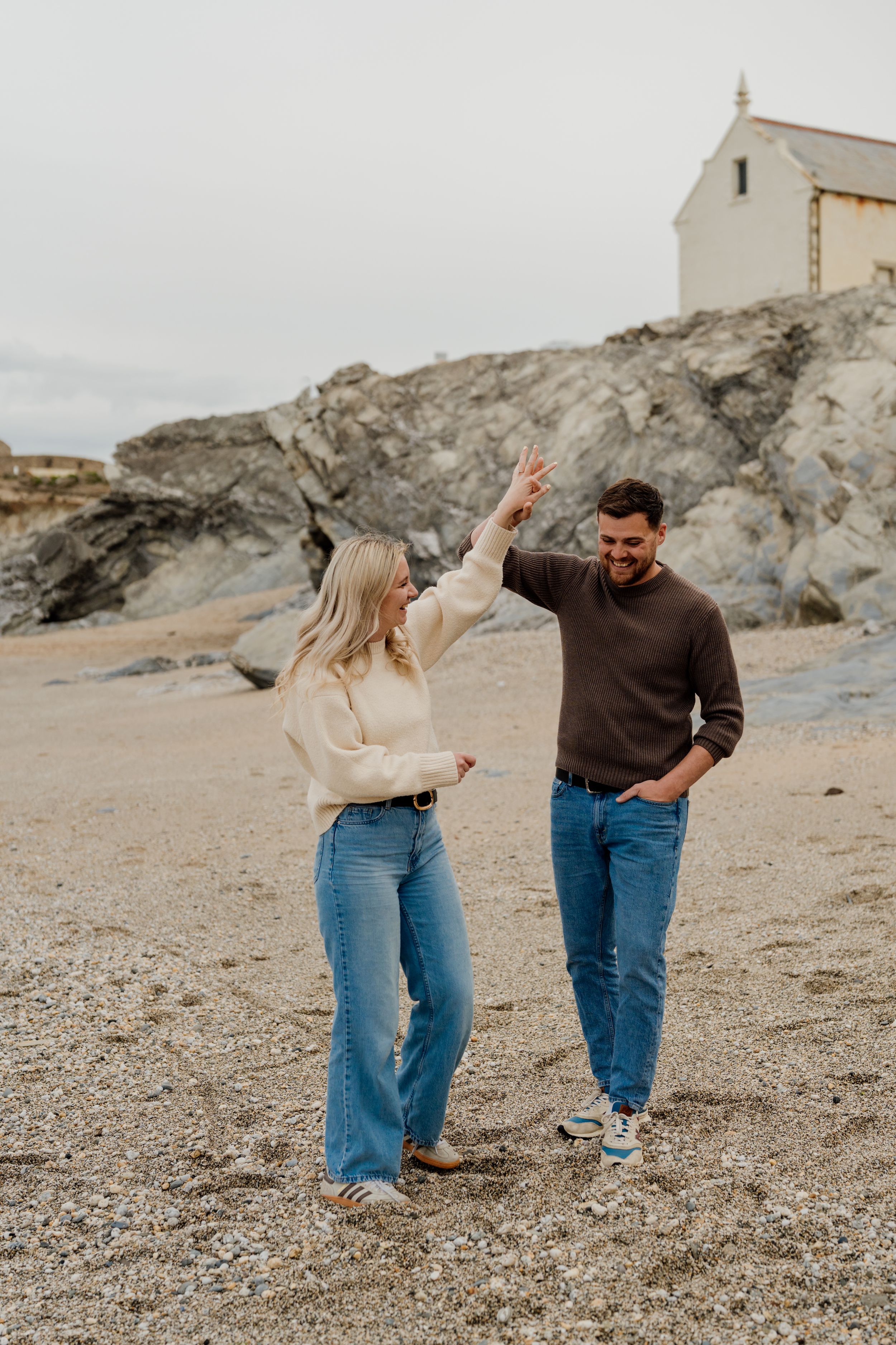 A couple dances on a rocky beach, the woman in a cream sweater and jeans, the man in brown, with a white chapel behind them.