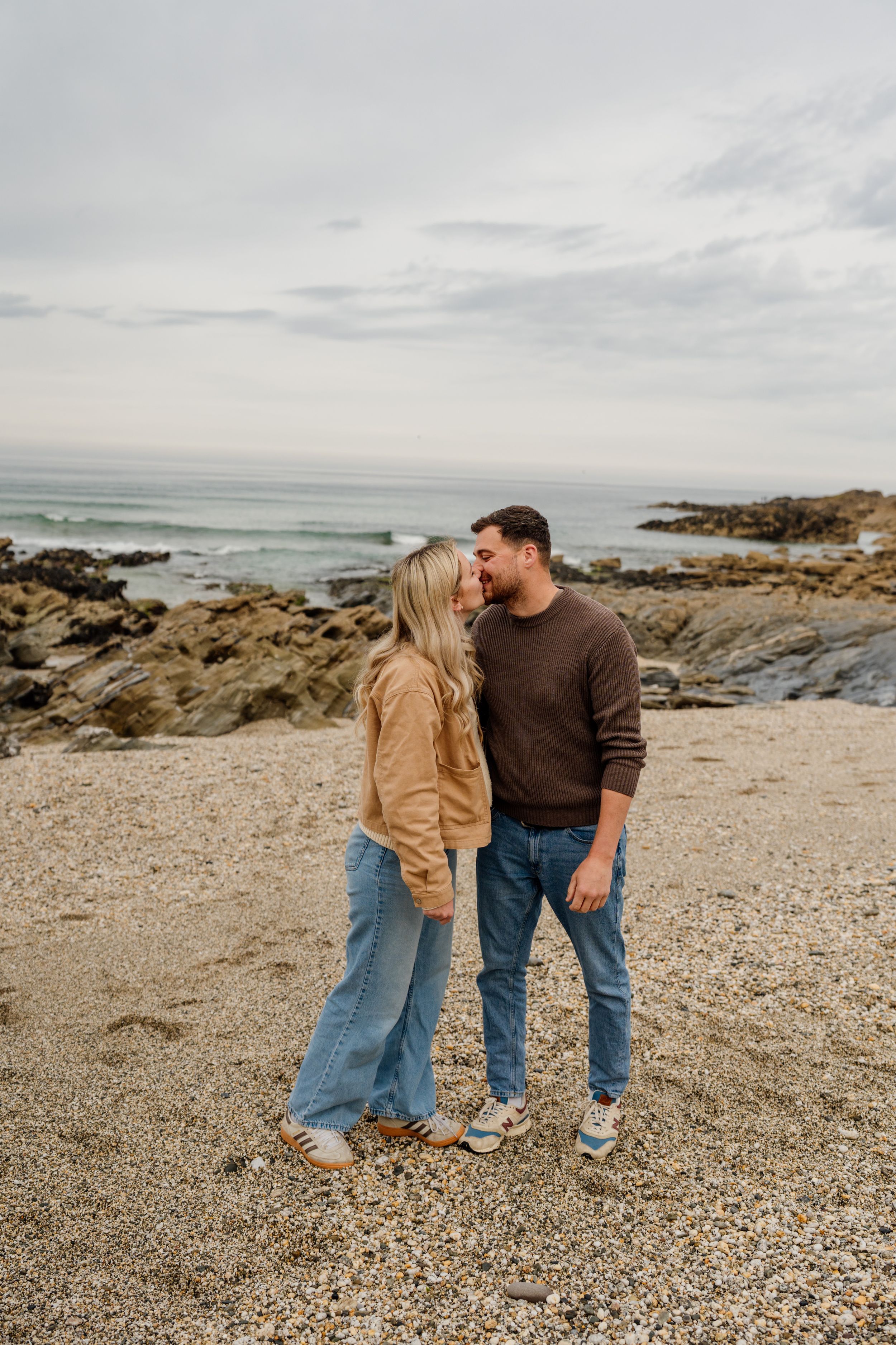 Couple sharing a romantic kiss on a rocky beach, wearing casual jeans and sweaters with ocean waves in the background.