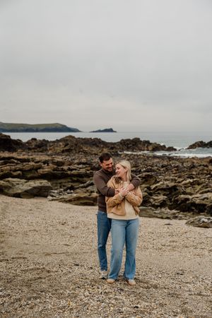 Couple embracing on a rocky beach with ocean views, wearing casual sweaters and jeans on an overcast day.